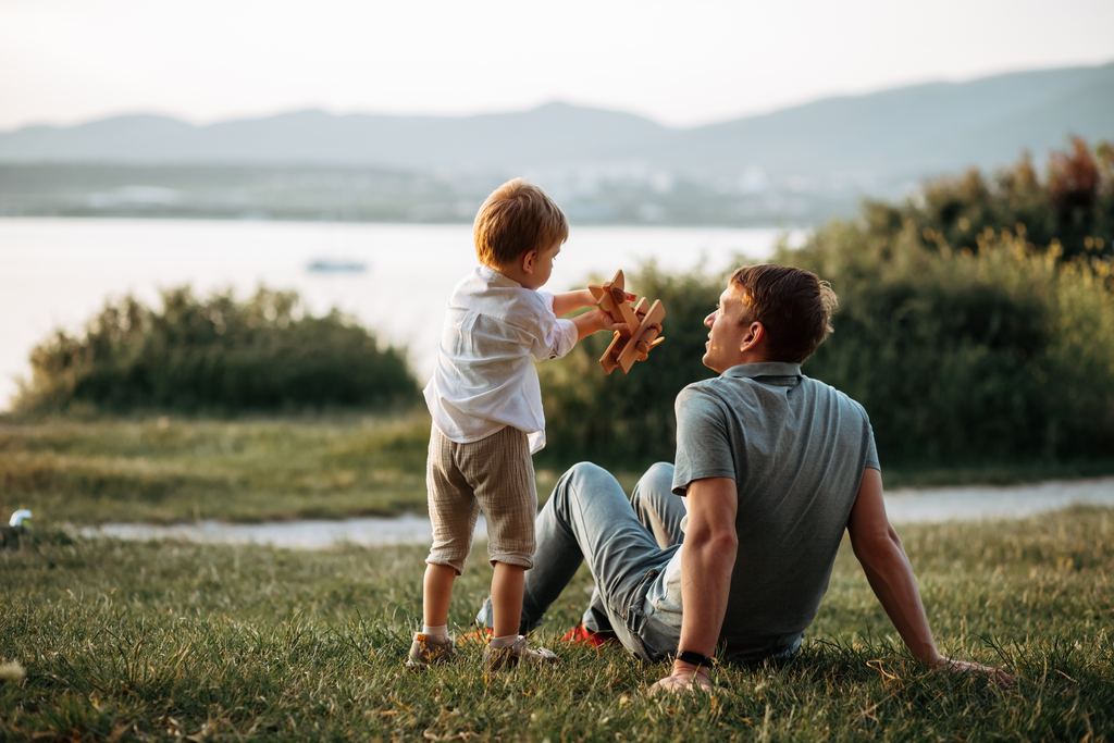 Un padre sentado en el pasto junto a un lago, mirando a su hijo pequeño que sostiene en alto un avión de juguete de madera.